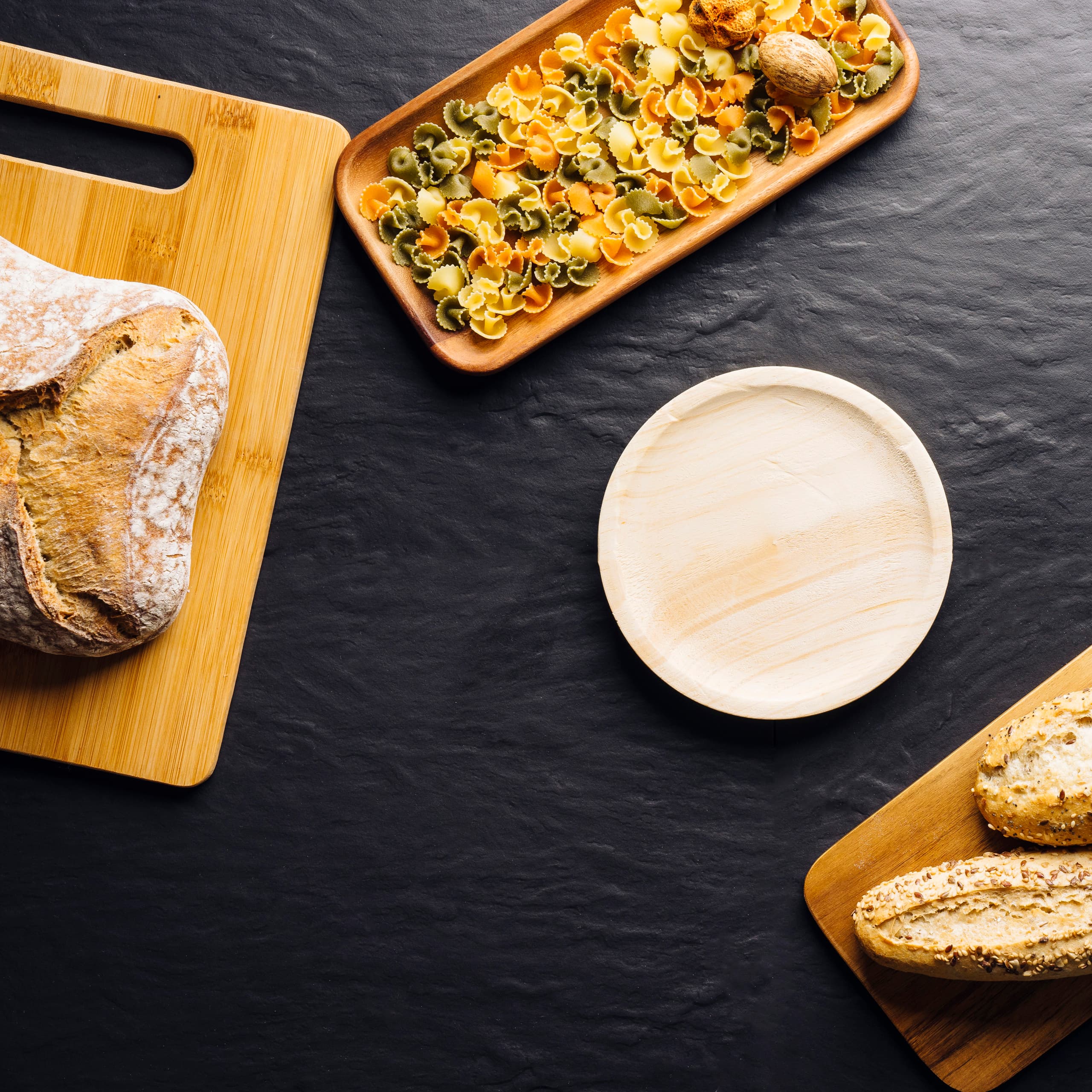 An empty wooden plate on a slate table, set with bread, boards and pasta